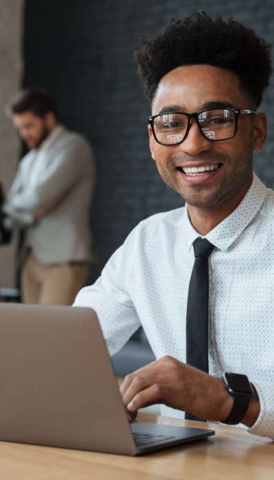 Photo of happy young african businessman sitting indoors using laptop computer. Looking camera.